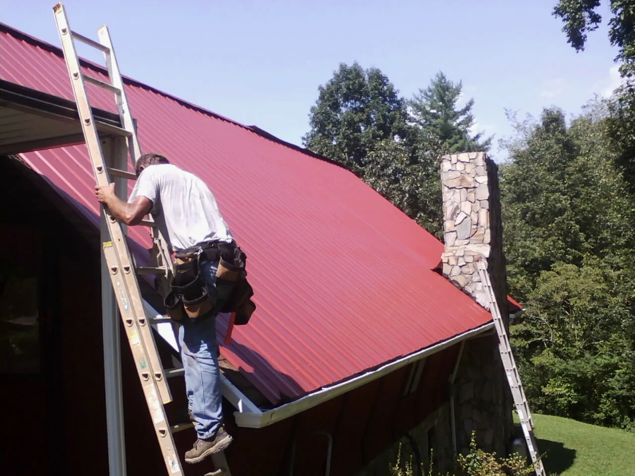 Roofer on a ladder beside a red metal roof and stone chimney — Soffit & Fascia Repair by Roofing Brothers in Refton